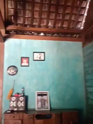 The video shows an interior view of a room with a tiled roof and wooden beams. The walls are painted turquoise. On the wall, there are three framed pictures, one of which is a circular clock. Below the pictures, there is a wooden cabinet with speakers and other objects on top, including a feather duster. To the right of the cabinet, there is another framed picture. In the background, through an open doorway, another part of the interior is visible, with furniture and a standing fan. The lighting appears to be natural light coming from the open doorway.