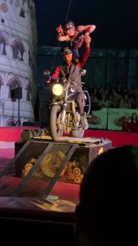 A performance is taking place under a circus tent, featuring a motorcycle stunt. A man dressed in a steampunk-inspired outfit, including a vest, red shirt, goggles, and hat, is seated on a motorcycle.  A woman, also in steampunk attire, is standing on his shoulders. The motorcycle is positioned on a small, elevated platform with ramps decorated with gold-colored gears. Behind them, there is a stage decoration depicting the Leaning Tower of Pisa.  A crowd of spectators can be seen in the background. The lighting is dramatic, with spotlights highlighting the performers and stage.