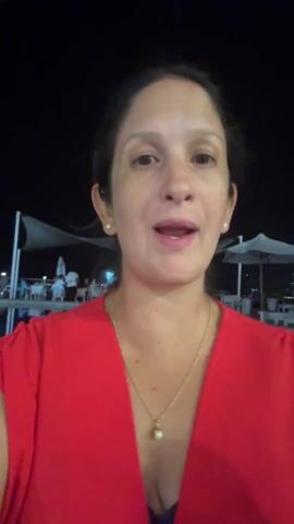 A woman in a red blouse and gold necklace stands in front of a pool at night. There are people and umbrellas in the background. She points to herself and adjusts her hair. The scene is dimly lit, and music is playing.
