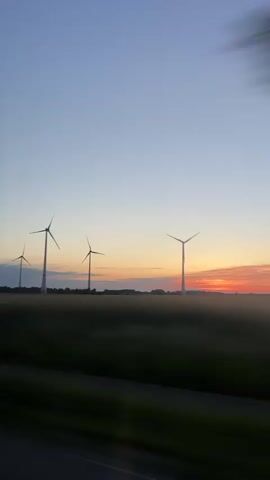 A landscape view shows four windmills standing on a field at sunset. The sky transitions from blue at the top to shades of orange and red near the horizon. Trees blur in the foreground, suggesting motion, likely filmed from a moving vehicle. The windmills are spaced across the horizon, providing a sense of scale. The foreground shows a stretch of green field.