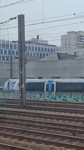A train moves through a station. The train is white and blue with graffiti on the side. In the background there are buildings and power lines. The tracks are in the foreground.