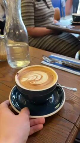 The video shows a hand holding a dark blue cup and saucer with a cappuccino on top. The cappuccino has a latte art design of what looks like leaves. Next to the cup are cutlery wrapped in a blue napkin, a glass jar filled with water, and a sugar shaker. The table is wooden. In the background, a person is sitting on a chair.