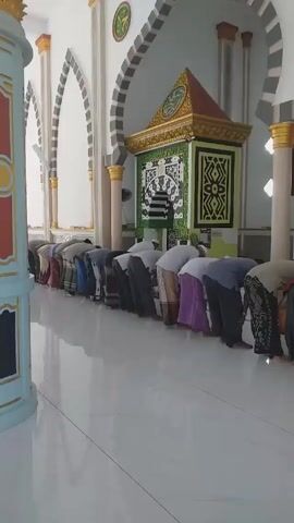 The video shows a group of men praying inside a mosque. They are lined up in a row, facing forward, and performing the bowing posture of the prayer. The imam, dressed in white, stands in front of them leading the prayer. The mosque has white tiled floors and arched doorways along the walls. There is a decorated niche (mihrab) on the wall, indicating the direction of prayer. The mosque appears to be well-lit and clean.