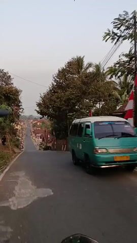 The video shows a person riding on a motorcycle down a paved road. The road curves to the left and goes downhill, with houses and trees on either side. There are Indonesian flags visible on poles along the side of the road. A green van is driving in the same direction as the motorcycle. The sky is overcast and appears to be early morning or late afternoon.