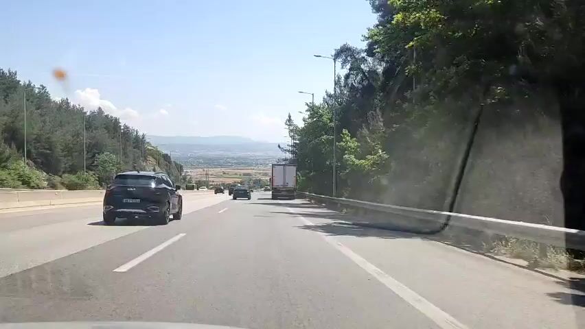 A dashcam view from a moving car shows a multilane highway under a clear sky. A black SUV drives in the left lane, and a white truck in the right. Other vehicles are visible in the distance. Hillsides with sparse trees line both sides of the road.