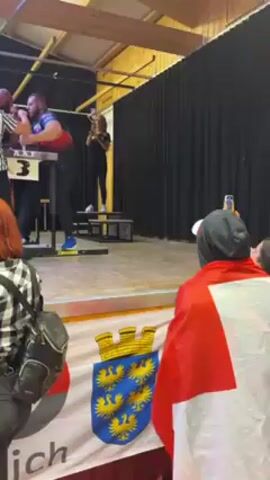 The video shows an arm wrestling match. Two men are competing on a raised stage with a black curtain backdrop. A female referee in a black and white striped shirt supervises the match. Another woman stands on a small platform to the side. A crowd watches the match from below, with one person draped in a Swiss flag and recording the event on a phone. The stage is decorated with a banner bearing a coat of arms and the word "ZUGO". The competition area is well lit, with visible beams and a simple ceiling structure overhead.