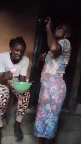 Two women are in the video. One woman is sitting on a small wooden stool, wearing a light gray t-shirt and leopard print pants. She is eating from a green bowl with her right hand. The other woman is standing, wearing a floral-patterned dress. She takes a drink from a dark bottle, then wipes her mouth with her hand. The background is a simple wall, and there's a dark doorway behind the standing woman.