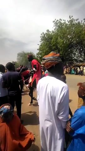 The video shows a group of people observing a horse parade or event. Several individuals in traditional clothing are visible, including a man in a white robe and turban in the foreground. Others wear colorful robes and headdresses. Some people are on horseback and wearing red and black attire. A crowd of onlookers stands in the background, suggesting a public event. The setting appears to be an open area with trees and a partly cloudy sky.