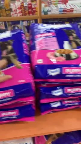 The video shows a person's hands reaching for bags of pet food on a shelf in what appears to be a store. The bags are stacked and labeled "Puppy" and have images of dogs on them. The shelves behind the pet food are stocked with various boxes and packages, some of which appear to be medicine or health products. The lighting is artificial, suggesting an indoor environment.