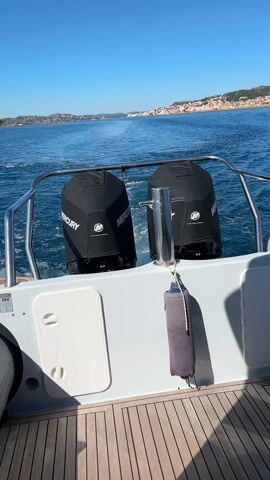 The video shows the back of a boat moving through the water, leaving a white wake. Two black Mercury outboard motors, covered with protective sleeves, are mounted on the boat's stern. A gray bumper is hanging from the railing. The deck is made of wooden planks. In the distance, land is visible under a clear blue sky. The sea appears calm, and the weather is sunny.