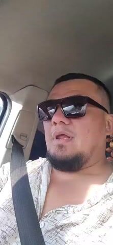 A man wearing sunglasses and a patterned shirt is in the driver's seat of a car, speaking. He has a goatee and a beaded necklace hanging from his right ear. The car appears to be in motion, with a view of a hilly, wooded landscape visible through the passenger window. The man's mouth is open as if he is speaking or exclaiming something.