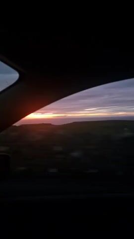The video shows a view from inside a vehicle looking out the window at a sunset over a body of water and rolling hills. The sky is a mix of purple, pink, and orange hues. The sun is a bright orange glow on the horizon. The landscape outside the window is blurred due to the movement of the vehicle, but appears to be rocky and sparsely vegetated. The inside of the vehicle is dark, with the dashboard and part of the steering wheel visible in the foreground. A side mirror is also visible on the left side of the frame. The overall mood is peaceful and serene.