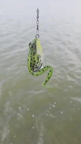 A fishing lure shaped like a frog hangs from a fishing line over rippling water. The lure is green and yellow with black spots and has a realistic frog appearance, including its legs and eyes. It is suspended by a swivel and a clip attached to the fishing line, which is barely visible against the muted background. The water is a murky green and appears to be calm with small ripples and a few white bubbles scattered on the surface. The lighting suggests it is daytime, possibly overcast.