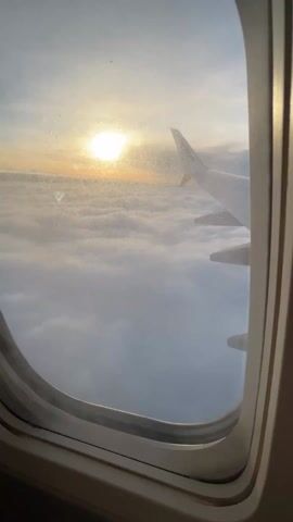 A video captured from inside an airplane shows a view out of the window. The sun, a bright circle, is visible low on the horizon, casting a warm orange glow across the sky above a thick layer of white and grey clouds. Below the sun, the clouds stretch out, resembling a vast, soft blanket.

The wing of the airplane is visible on the right side of the frame, extending into the cloudscape. The winglet, the upward-curved tip of the wing, is clearly discernible, and the word "Ryanair" is faintly visible on it. The airplane's engine is partially visible below the wing. The window frame itself takes up the immediate foreground, slightly dirty with smudges, adding to the realism of the aerial journey.

In the background, a person can be heard speaking in Polish, making an announcement. The overall impression is one of peaceful flight during either sunrise or sunset.