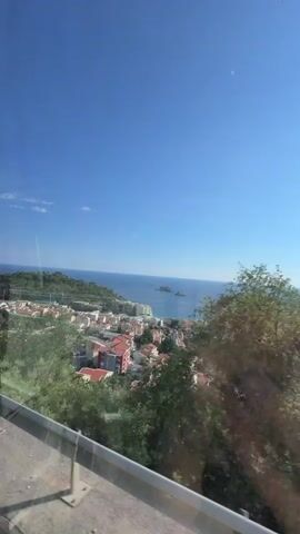 This media file captures a scenic view of a coastal town in Stupovi, Montenegro, on a bright, sunny day. The scene looks out over a cluster of buildings with red-tiled roofs, interspersed with patches of green trees and lush vegetation. In the distance, the deep blue sea stretches to the horizon, dotted with a few small islands. The sky is a clear, vibrant blue, with a few wispy white clouds.

The perspective suggests the shot was taken from an elevated position, possibly from a vehicle or a hillside overlooking the town. A metal guardrail runs along the bottom of the frame, indicating a pathway or road. The time of day appears to be late morning or early afternoon, given the bright sunlight casting clear shadows. The overall mood is one of tranquility and scenic beauty, typical of a Mediterranean coastal landscape. No people or text are visible in the frame.