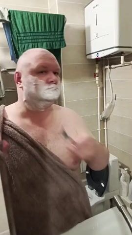 A man shaves his face with a razor. He is wearing a towel draped over his shoulder. The background appears to be a bathroom with a tiled wall and a water heater.