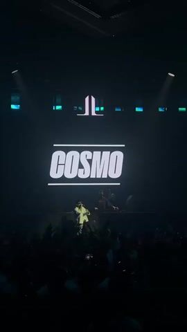 A dark concert venue is illuminated by stage lights. A large screen behind the performers displays the word COSMO in bold white letters. In front of the screen, on a raised platform, a singer wearing a white t-shirt and shorts is on stage. To his right, two DJs stand behind a console. A large crowd of people is visible in the foreground, facing the stage, with many holding up their hands and some holding up cell phones. The lighting is dramatic, with spotlights focused on the performers and the crowd. The overall atmosphere is energetic and celebratory.