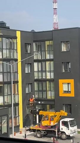 This media file shows an exterior view of a modern apartment building. The building features a dark gray facade with bright yellow accents and numerous windows. A construction scene is in progress, with a yellow crane truck parked in front of the building. A lift basket is extended up the side of the building, carrying two construction workers wearing orange safety vests. One worker appears to be working on the exterior of the building near a window, while the other stands in the basket. In the background, a tall, red and white radio tower is visible against a clear sky, suggesting fair weather. The time of day appears to be daytime, given the lighting. The scene suggests ongoing construction or maintenance work on the apartment complex. There is no visible text in the media file.