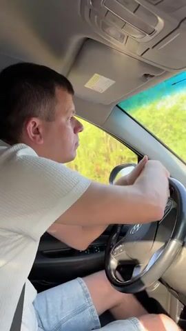 A man drives a car. The camera view begins looking at the driver and the steering wheel, then switches to look out of the front windshield. The car is approaching an overpass with a height restriction sign of 3.3 meters. Green vegetation is visible beyond the bridge. The car continues to move forward, passing beneath the overpass. The time on the car’s dashboard display reads 07:58, and the radio station is set to 95.8 FM. A dashcam is affixed to the windshield behind the rearview mirror. The man is wearing a light-colored, short-sleeved shirt and light blue shorts.