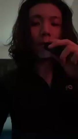 A young person with long dark hair is holding a black vape pen up to their mouth and exhaling. They are wearing a black shirt with a small white logo on the right breast. The room is dimly lit, with a red light illuminating the person's face. There is an air conditioner on the wall in the background. Music is playing in the background.