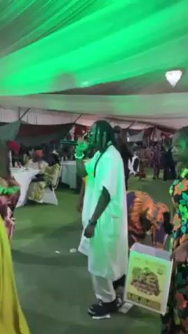 The video shows a live performance at an event. A man in a white robe is singing into a microphone while dancing. He has dreadlocks and sunglasses. Several women are dancing around him. One woman is wearing a gold and green dress, and another is in a floral print dress holding a small box with money in it. The venue appears to be a tent or covered area with green lighting. Other people are visible in the background, sitting at tables.