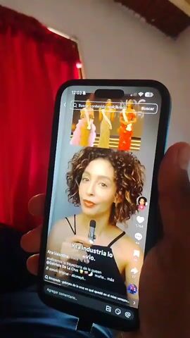 A hand holds a smartphone showing a TikTok video featuring a woman in a red dress and a "Miss Grand View All Stars" sash. She is wearing a crown and standing in front of the Venezuelan flag and a seal. The video's caption reads, "Meses después recibió esa llamada, sin avisarle porque Venezuela necesitaba una." The TikTok interface includes like, comment, and share icons. The background of the scene shows a red curtain.