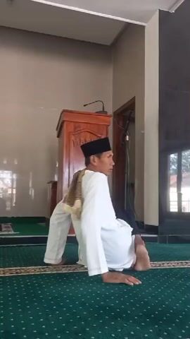 A man in a white robe and black pants, wearing a black skullcap and a patterned sash, is seen performing a sequence of movements inside a mosque. He is barefoot and on a green carpet with a repeating diamond pattern. In the background, a wooden podium with carvings and a microphone is visible, as are parts of the mosque's interior, including walls and a doorway. The man begins in a low, seated position on the floor, then rises to a standing posture, touching his stomach. He then walks a few steps to the right.
