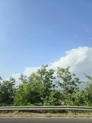 The video shows a view from a moving vehicle, looking towards a bright blue sky with scattered clouds. In the foreground, there is a metal guardrail. Beyond the guardrail, there is a flat expanse of land that appears to be agricultural, with some structures and vegetation. Further in the distance, a large, modern sound barrier made of metal and transparent panels is visible. The sound barrier has several bird-like silhouettes affixed to it. The video is shot from a low angle, suggesting the vehicle is on a road.
