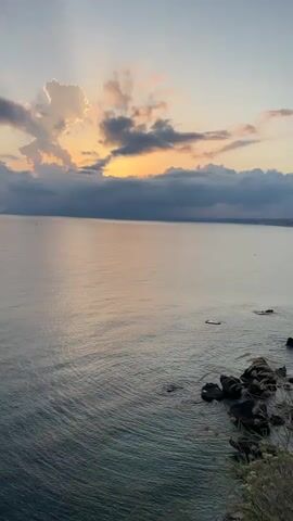 The video displays a tranquil ocean at sunset. The sky is painted with soft hues of orange, pink, and yellow, peaking through a scattering of clouds. The ocean's surface reflects these colors, creating a serene and peaceful atmosphere. Jagged rocks are visible along the coastline in the foreground, with some vegetation growing near them. The water is calm, with gentle ripples. A small boat is barely visible in the distance. The overall impression is one of natural beauty and quietude.