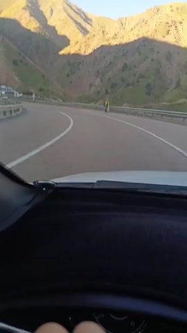 The video begins from the perspective of a passenger inside a vehicle, looking forward at a highway. Mountains with terraced slopes are visible in the background, bathed in the warm light of what appears to be sunrise or sunset. The road ahead curves gently to the left. The camera then pans to the right-side mirror, showing a reflection of the road and mountains, with the car's exterior visible. The view returns to the front of the car, now showing the driver's hands on the steering wheel. The highway continues forward, with several cars in the distance. The sun illuminates the mountain slopes, creating a dramatic contrast between light and shadow.