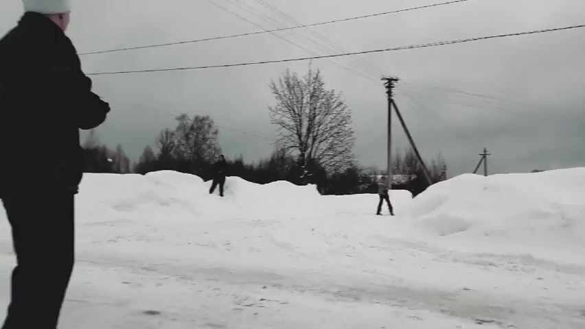 A man in a black jacket and white hat stands to the left of the frame, looking at two children playing in the snow-covered area. The children are near a snow bank, amidst a winter scene under an overcast sky with bare trees in the background and utility poles strung with lines. The man throws something, perhaps a snowball.