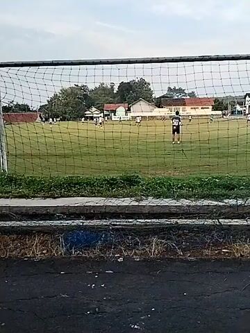 The video shows a soccer field viewed through a goal net. Players wearing white and blue uniforms are seen on the field. A player wearing a black uniform with the number 50 on the back stands near the center of the field, facing away from the camera. In the background, there are several buildings and trees. The sky is overcast. The ground in front of the net is asphalt with some grass and debris along the edge.