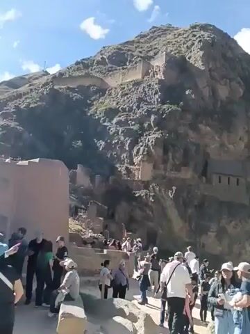 The video shows a mountainous landscape with ancient ruins and a crowd of people exploring the site. The camera pans across the landscape to reveal the mountainous terrain, with some trees growing on the mountain sides, and a village at the foot of one mountain. There are also some structures built into the side of one of the mountains. A large group of people is standing in the foreground, examining the ruins. Some people are standing still while others are walking around. The sky is blue with scattered white clouds. The overall impression is that of a historical site in a mountainous setting, with many tourists visiting to explore.