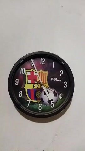 A close-up shot of a round clock with a black frame is mounted on a white wall. The clock face features the FC Barcelona crest and a soccer ball design. The clock hands are visible, indicating a time around 10:15. The video captures a slight wobble of the clock, suggesting it might be attached loosely or in a slight breeze. The background is a plain white wall, offering no additional context.