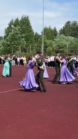 On a red outdoor basketball court with a tall metal light pole, multiple couples, each a woman in a long, flowing lilac dress and a man in a dark green military uniform with a gold sash, dance together. Several other couples are also present, including a woman in a light green dress. The dancers are surrounded by a large group of spectators standing behind a green chain-link fence, beyond which is a line of trees. The sky is bright blue, with scattered white clouds. The camera angle is mid-level, and the perspective shifts slightly as the dancers move.