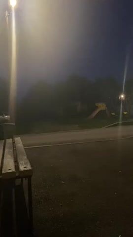 The video opens with a view of a park or playground at dusk or night. The lighting is dim, with streetlights casting a glow and creating lens flares. In the foreground, the edge of a wooden bench is visible on the left. A paved area, likely a path or road, runs horizontally across the middle ground. On the right side of the paved area, a playground structure with a red and yellow slide and swings is faintly visible, partially obscured by fog or mist. Trees and other structures are silhouetted in the background. The overall atmosphere is dark and slightly hazy. The camera appears to be stationary.