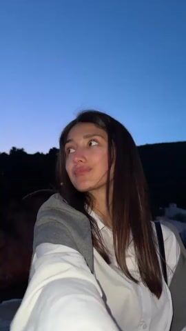 A woman with long, straight brown hair is standing outside. She is wearing a white button-down shirt and a gray sweater draped over her shoulders. She is looking at the camera, and she winks. Behind her, there are silhouetted trees and what appears to be a building. The sky is a gradient of blue.