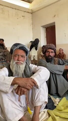 The video shows a group of men sitting on the floor inside a room. The room appears to be sparsely furnished with bare, cream-colored walls and a corrugated metal roof. Several men are wearing traditional clothing, including turbans and long robes. One man in the foreground, with a long white beard, is looking directly at the camera. He is wearing a white robe and a grey turban. Another man, with a dark beard and turban, is sitting to his right and looking away from the camera. In the background, several other men are visible, some sitting and some standing. One man in the background appears to be a young boy. The lighting in the room is natural, coming from above and from an open doorway. The overall impression is of a gathering or meeting taking place in a simple, rural setting.