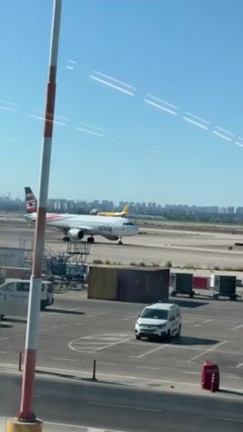 The video shows an airport tarmac on a sunny day. In the foreground, a white van with "ARKIA" printed on its side is parked in a marked parking area. To the left, a striped pole, likely part of an airport fence or barrier, is visible. In the middle ground, a white airplane with red and black accents on its tail, and the word "ARKIA" on its fuselage, is on the runway, appearing to be taxiing. Further in the background, a yellow cargo plane with "DHL" on its side is also on the runway, moving slowly. A cityscape with buildings and construction cranes is visible in the far background under a clear blue sky. The overall scene depicts typical airport operations with aircraft and ground vehicles.