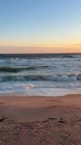 The video showcases a beach at sunset. The waves are crashing onto the sandy shore, creating a foamy white line along the edge. The sand itself is a mix of light brown and reddish hues. As the camera pans upward, it reveals the horizon where the sun is setting, casting a warm orange glow across the sky and reflecting on the water. The sky transitions from a light blue near the top of the frame to shades of orange and pink near the horizon. There are a few faint clouds in the distance, adding texture to the sky. A contrail is briefly visible in the sky.