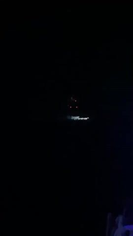 A fireworks display is occurring over a body of water at night. A small cluster of red fireworks is visible in the dark sky, with their reflection appearing on the water below. A distant boat or platform is illuminated by lights, possibly the source of the fireworks. The scene is dark with the exception of the fireworks and the boat's lights.
