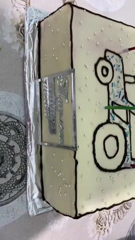 The video showcases a rectangular cake decorated with white frosting. The cake features a dark chocolate outline of a tractor, filled with blue sprinkles. Several small, pearl-like candies are scattered across the frosting. On one side of the cake, there is a silver metallic frame with text that appears to say "MARATHON". Red and green wires or ribbons are inserted into the cake, along with a few unlit candles. The cake is placed on a table covered with a tablecloth featuring a circular, mandala-like pattern in shades of gray and white. The lighting suggests it is daytime, and the setting appears to be indoors, possibly a home. There are no people visible in the video. The overall impression is of a celebratory cake, likely for a birthday or special occasion. The location context provided is Myrza-Ake, Kyrgyzstan, which adds a regional cultural element to the scene.