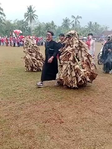 The video shows a parade or outdoor event with several people walking across a grassy field. In the foreground, a person in a costume made of dried leaves walks towards the left. To their right, a person dressed in black with sunglasses walks forward. Further to the right, a person with white and red body paint walks. Behind them, a person in a dark, shaggy costume walks. In the background, there are more people, some riding motorcycles, and several clusters of pink and white balloons floating in the air. There are trees and tents visible in the distance, suggesting a festive outdoor gathering.