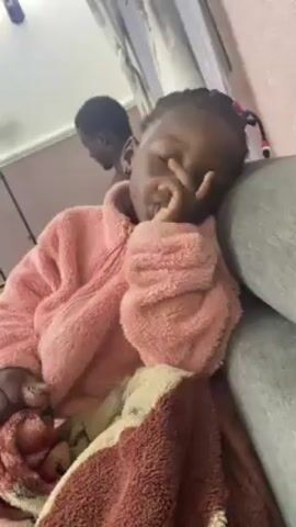A young Black child wearing a pink fuzzy jacket is sitting on a gray surface with a brown and tan patterned blanket. The child is holding a finger to their mouth, possibly falling asleep. Another child is visible in the background. Someone off-screen says "Say Happy New Year" and "You not say Happy New Year?"