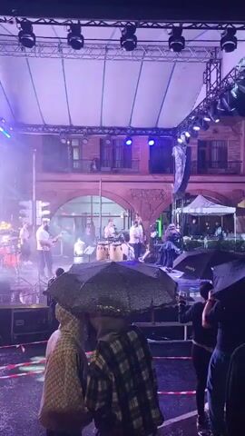 The video shows an outdoor concert scene at night. The stage is illuminated with blue and purple lights, and a band is performing, with instruments visible like guitars and drums. Several people are standing in the foreground, holding umbrellas, suggesting it might be raining. The background reveals buildings and a street scene with more people gathered, some also holding umbrellas. A pedestrian crossing sign and a traffic sign are also visible. The overall atmosphere appears to be of a public event in progress despite the weather.