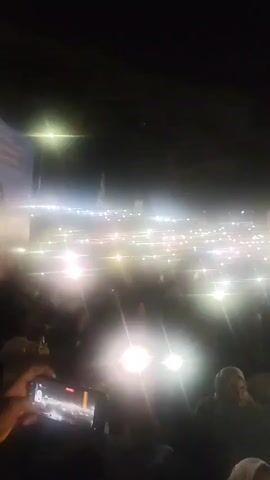 A low-angle, handheld shot captures a nighttime outdoor gathering. The scene is dominated by a dense crowd of people, mostly seen from the chest up. Numerous individuals are holding up their mobile phone flashlights, creating a widespread, bright illumination across the scene and casting lens flares. In the background, there is a large, tall, vertical structure with yellow and green bands, possibly a flagpole or monument. The overall atmosphere suggests an event or demonstration, with the flashing lights indicating collective engagement and potential recording of the proceedings. The environment appears dark with the exception of the artificial lights.