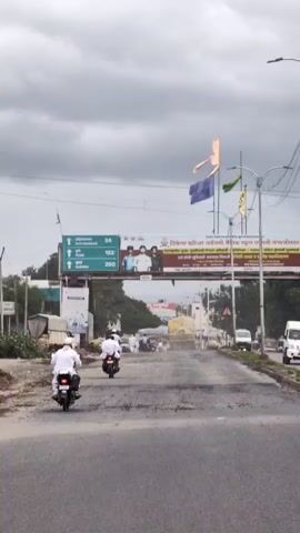 A vertical video shows a street scene under a cloudy sky. In the foreground, a person on a white motorcycle with a black seat rides away from the camera on a paved road with visible potholes. Another person on a similar motorcycle is ahead of them, also moving away. In the background, a few other vehicles are visible on the road, including a truck with a large yellow tarped load. A large overhead banner spans the road, featuring advertisements and text in an Indian language. On the left side of the banner, there is a green section with white text and numbers, possibly indicating distances to cities. The banner also displays images of several people. Above the banner, two tall poles with flags are visible: one with a yellow and orange flag, and another with a blue flag. The overall impression is a busy street in a town or city, likely in India, given the signage and vehicles.