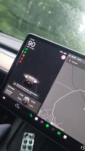 The video shows the interior of a car, focusing primarily on the central touchscreen display and then transitioning to the view through the windshield. The scene depicts a vehicle being driven during heavy rain.

Inside the car, a Tesla-style touchscreen dominates the view. At the top left of the screen, the speed is displayed as 89 km/h (55 mph), then briefly 88 km/h. Several warning icons are visible, including a red warning triangle, an orange airbag icon, and a yellow tire pressure icon. The screen shows two explicit warnings: "Safety restraint system issue - Service required. Adult occupants may not be restrained." and "Tire Pressure: Check tire pressure. Your tire pressure is low. See owner's manual for details." Detailed tire pressure readings are displayed: front left 3.3 bar, front right 3.4 bar, rear left 2.3 bar, and rear right 2.3 bar. The map on the right side of the screen shows a road network with a red destination marker, and the text "Orthodox church" is faintly visible. The time is 08:08, the outside temperature is 18°C, and the battery charge is 82%. A circular speed limit sign of "50" is also present. Below the screen, partially visible on what appears to be a passenger seat or console, is a white blister pack containing several green, round pills.

The view outside the windshield shows a two-lane road lined with dense green trees. Heavy rain is falling, creating numerous droplets on the windshield and making the road surface wet and reflective. Headlights from an oncoming vehicle are visible in the distance. Guardrails line the sides of the road. The overall impression is of driving through a forested area in rainy, overcast conditions. The location context provided is Borovtsi, Bulgaria.