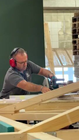 A man wearing a grey polo shirt, glasses, and red ear protection is using a circular saw to cut a piece of wood. The wood is part of a larger wooden structure, possibly a frame. He is focused on the task, holding the saw with both hands. The background shows a large green wall on the left and a window with wooden pallets stacked to the right. The lighting suggests an indoor environment, possibly a workshop or construction site.