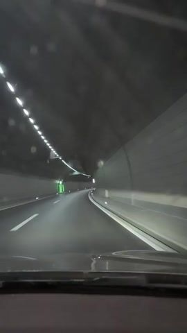 The video shows the interior of a car driving through a tunnel. The perspective is from the driver's seat, looking out the windshield. The tunnel is dark with overhead lighting that is illuminated in a dashed pattern. The road ahead curves to the right. There are white lane markings on the road. A green sign with white lettering indicating "Lausanne" is visible on the left side of the tunnel. The car appears to be moving at a moderate speed. The overall environment is that of a modern, well-lit highway tunnel.